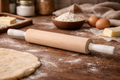 Wooden rolling pin on a wooden surface with flour, eggs, and other baking ingredients.