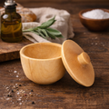 Wooden bowl with lid on a wooden surface with herbs and oil in the background