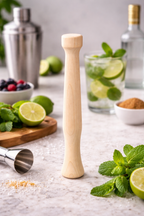 Wooden muddler on a table with limes, mint, and cocktail ingredients.