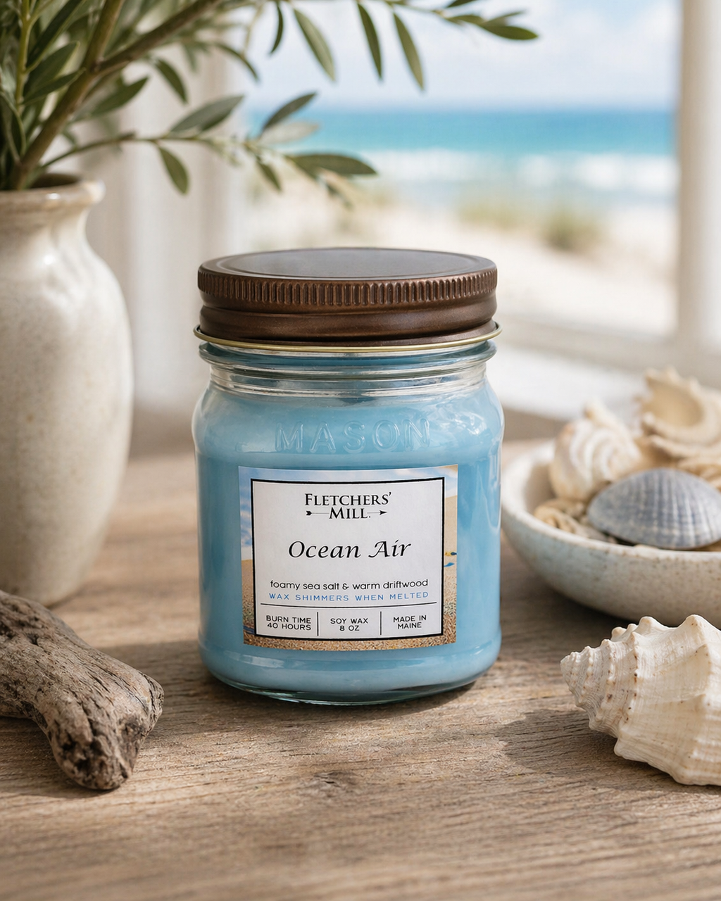 Candle labeled 'Ocean Air' by Fletcher's Mill on a wooden surface with seashells and a plant in the background.