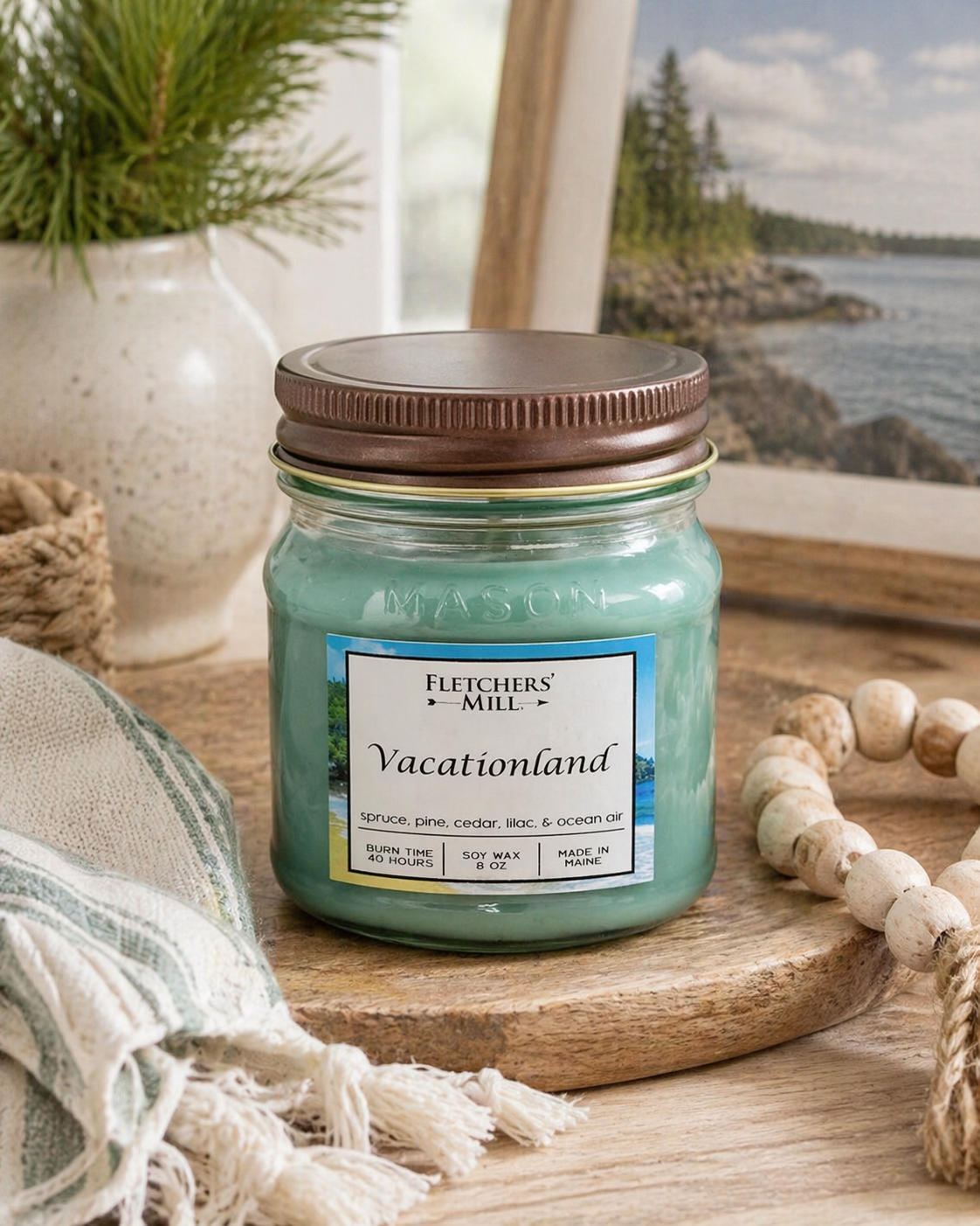 Jar candle labeled 'Vacationland' by Fletchers Mill on a wooden surface with a scenic background.